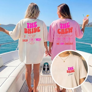 Può includere: Due donne indossano magliette oversize con testo. Una maglietta è color crema con "THE BRIDE" e "I SAID YES!" stampato sul retro. L'altra è rosa con "THE CREW" e "WE SAY CHEERS!" sul retro. Un'immagine più piccola mostra "THE BRIDE" e un nome.