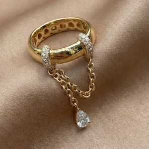 May include: A gold ring with a unique design. The ring features a thick band with small circular cutouts and two sections with small clear stones. A delicate gold chain hangs from the ring, ending in a pear-shaped clear stone.
