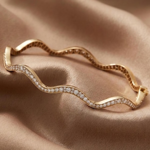 May include: A gold-toned, wavy bracelet with a continuous line of small, sparkling gemstones. The bracelet is set against a soft, satin-like background, highlighting its elegant design and craftsmanship. The bracelet's design is reminiscent of ocean waves.