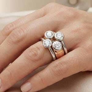 May include: A multi-band ring featuring four round, bezel-set clear stones. The design incorporates a combination of silver-toned and rose gold-toned metal bands, creating a layered appearance with the stones clustered together on the finger.