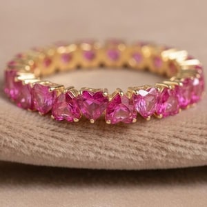 May include: A gold ring with a continuous row of heart-shaped pink gemstones. The ring's band is polished gold, and the gemstones are set closely together, creating a full circle of vibrant colour. The ring is displayed on a neutral, textured surface.
