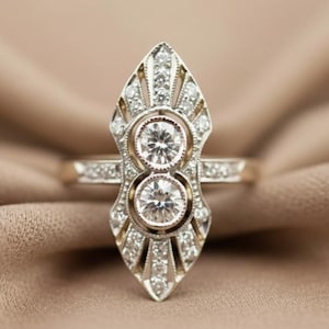 May include: An Art Deco style ring with two prominent round diamonds set in a decorative, elongated setting. The ring features intricate detailing with smaller accent diamonds and a gold band. The design is symmetrical and elegant, perfect for a special occasion.