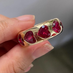 May include: A gold band ring featuring a series of faceted, heart-shaped red stones set into the metal. The ring has a smooth, polished finish and the stones are arranged horizontally around the outer surface of the band, creating a continuous pattern.