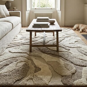 May include: A large, textured area rug with a flowing, abstract design in shades of beige and brown. The rug is placed in a living room setting, with a sofa, coffee table, and fireplace visible in the background. The rug's dimensions are approximately 8 x 10 feet.