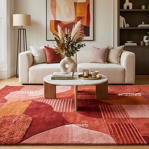 May include: A modern living room featuring a large, abstract patterned rug in shades of red, orange, and pink. The rug is the focal point, complemented by a cream-colored sofa, a round coffee table, and an orange armchair. Decorative elements include pillows, a vase with dried flowers, and artwork.