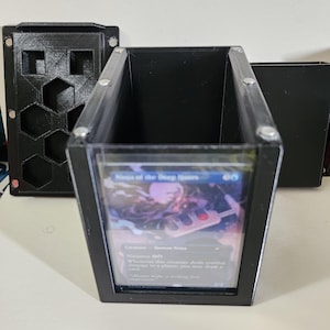 May include: Black rectangular card holder with a clear front panel, holding a Magic: The Gathering card. The holder is made of black plastic and has silver rivets. A separate black lid and a black card holder with hexagonal cutouts are also visible.