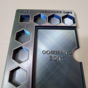 May include: A metallic grey game card holder. It features the text "CASTS COMMANDER DMG", "LIFE", and "COMMAND ZONE". The holder has various shaped slots and a rectangular card slot.