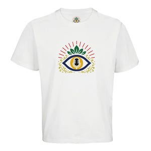 Puede incluir: Camiseta blanca con un diseño de ojo colorido que incorpora un micrófono. El ojo está delineado en azul y dorado, con detalles rojos y verdes. Una versión más pequeña del diseño está en el cuello.