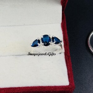 Può includere: Un anello in argento con una gemma blu scuro taglio ovale al centro, affiancata da due gemme blu scuro più piccole taglio a goccia. Il testo InayajewelsGifts è stampato in nero sotto l'anello su uno sfondo bianco.