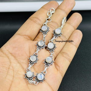 Könnte beinhalten: Ein silbernes Armband mit Mondstein-Cabochons in einem floralen Design. Das Armband hat runde, milchig-weiße Mondsteine, die jeweils in einer detaillierten, sternförmigen Fassung gefasst sind. Der Verschluss ist sichtbar, und der Text "Inayajewels Gifts" ist vorhanden.
