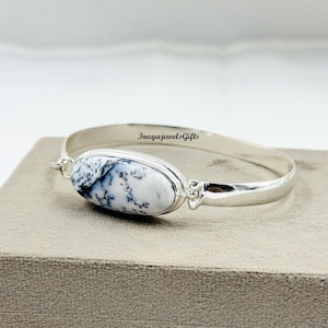May include: A silver bangle bracelet with a white and black dendritic agate stone. The oval-shaped stone is the focal point, set in a silver bezel. The bracelet is on a neutral surface. The text "InayajewelsGifts" is on the bracelet.