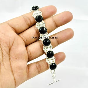 May include: A silver bracelet featuring six large, round, black gemstones. The bracelet has a toggle clasp and the text "INAYAHJEWELSGIFTS" is visible. The bracelet is held in a hand.