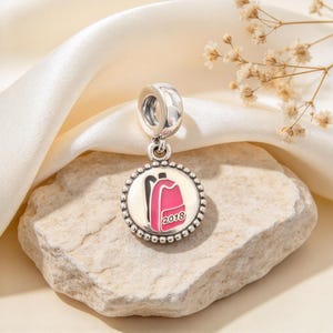 Puede incluir: Un charm de plata con un colgante circular de borde perlado. El centro muestra un gráfico de mochila rosa con contornos negros y el texto 2018 impreso en negro. El charm tiene una anilla de plata pulida para sujetar a una pulsera.