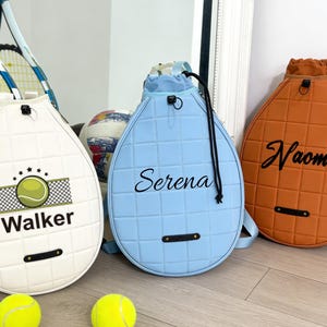 May include: Three tennis racket-shaped drawstring bags in white, light blue, and orange. The white bag has a tennis ball graphic and the name "Walker". The light blue bag says "Serena", and the orange bag says "Naomi". Two yellow tennis balls are in the foreground.