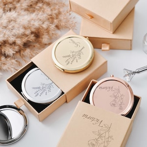 以下が含まれることがあります： 金、銀、ローズゴールドのパーソナライズされたコンパクトミラーのコレクション。それぞれに名前と花の模様が刻まれています。ミラーは、開いたベージュのギフトボックスに表示されています。透明な化粧品ツールが見えます。