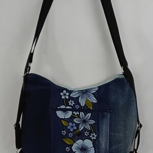 May include: A denim shoulder bag with a floral design. The bag has a dark blue denim base, a light blue zip, and a black adjustable strap. The front is decorated with embroidered blue flowers and green leaves.