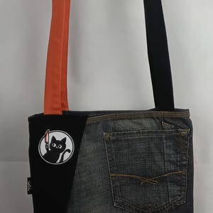 May include: A denim and black fabric tote bag with a black cat graphic holding a knife. The bag has orange and black straps, and patches with the text "IT IS WHAT IT IS" and "THAT'S A TERRIBLE IDEA WHEN DO WE START."