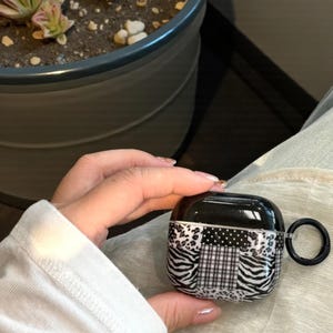 May include: Black and white patterned AirPod case with a black carabiner. The case features a patchwork design with zebra stripes, polka dots, and plaid patterns. The case is held in a person's hand.