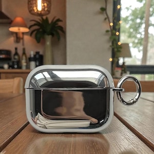 May include: A shiny, silver-colored AirPod case with a white border and a silver ring attachment. The case is sitting on a wooden table, reflecting the surrounding environment. The case has a smooth, polished surface.