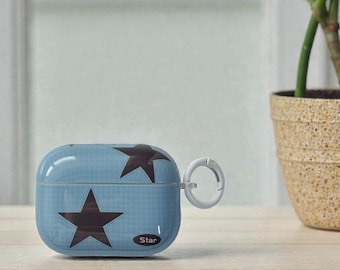 May include: A light blue AirPod case with a repeating star pattern in dark brown. The case has a white carabiner clip and the word "Star" printed on the bottom. The case is sitting on a light brown wooden surface.