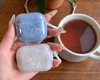 Może przedstawiać: Dwa etui na AirPods trzymane są w dłoni. Jedno jest jasnoniebieskie z kwiatowym wzorem, a drugie białe z podobnym wzorem. Filiżanka herbaty i roślina doniczkowa znajdują się w tle. Etui mają ochronny design.