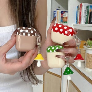Könnte beinhalten: Drei AirPod-Hüllen mit Pilzdesigns. Eine Hülle ist braun mit weißen Punkten, eine andere ist rot mit weißen Punkten und die dritte ist grün mit weißen Punkten. Jede Hülle hat einen passenden Pilz-Anhänger an einem Schlüsselanhänger.