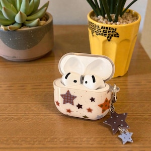 Könnte beinhalten: Ein weißes AirPod-Case mit braunen und orangefarbenen Sternen. Das Case ist geöffnet und zeigt weiße Ohrhörer. Ein sternförmiger Anhänger mit braunen und blauen Sternen hängt am Case. Es liegt auf einer Holzoberfläche.