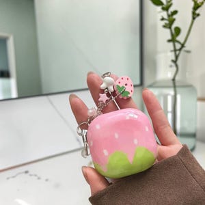 May include: A glossy, square-shaped protective case for wireless earbuds, featuring a pink base with white strawberry seed patterns and a green scalloped bottom. A silver-toned metal chain is attached, adorned with small charms including a strawberry.