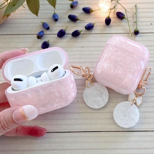 May include: Pink marbled AirPods Pro case with a matching open case containing white AirPods. A closed case and two white circular charms with gold-colored accents are also present. The background includes scattered berries and greenery.