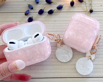 Może przedstawiać: Różowe, marmurkowe etui na AirPods Pro z pasującym otwartym etui zawierającym białe słuchawki AirPods. Obecne jest również zamknięte etui i dwie białe okrągłe zawieszki ze złotymi akcentami. Tło zawiera rozrzucone jagody i zieleń.