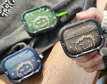 May include: Three transparent AirPod cases in green, blue, and gray, each featuring a white graphic of a stitched face. The cases have a hard shell design with a clear front and a colored frame. One case is held by a person.