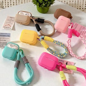 May include: Several brightly coloured AirPod cases in shades of tan, yellow, pink, and teal, each with a matching lanyard. Some cases are open, showing the white earbuds. Small cards with text are also visible in the image.