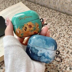 May include: Two teal and blue AirPod cases. One case features a sunflower design with the word "Sunflower" in gold script. The other case has a blue and white wave pattern. Both cases have a silver keychain attachment.