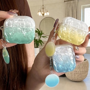 May include: Three clear, textured cases for wireless earbuds in shades of blue, yellow, and green. Each case has a matching keychain with a decorative charm. The cases are held up by a person.