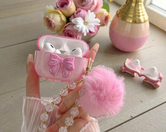 May include: A pink AirPod case with a bow design, embellished with rhinestones, and a fluffy pink pom-pom keychain. The case is held in a hand, with a clear beaded strap. The background includes a vase of flowers and a decorative perfume bottle.
