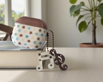 May include: A white and brown AirPod case with a star pattern, connected to a brown chain and a musical note charm. The charm features a white quarter note and a brown treble clef, both with star cutouts.