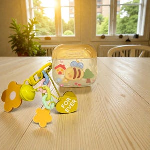 May include: A clear, square case with a yellow lid and a bee graphic. Attached is a yellow keychain with flower and heart-shaped charms. The heart charm reads "FOR EVER".