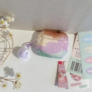May include: A clear AirPod case with a colourful design featuring shades of brown, white, purple, and green. The case has a small purple pom-pom attached. The image also includes small cards with text and a barcode.