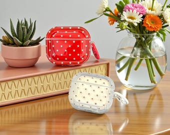 May include: Two AirPod cases with star patterns. One is red with white stars, the other is clear with tan stars. A small succulent in a pink pot and a vase of flowers are also in the image.
