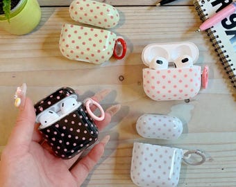 Può includere: Una collezione di custodie per AirPod in vari design. Una custodia è nera con pois rosa, un'altra è bianca con cuori rosa, e altre sono bianche con pois rosa. Ogni custodia è aperta, rivelando gli AirPods all'interno.