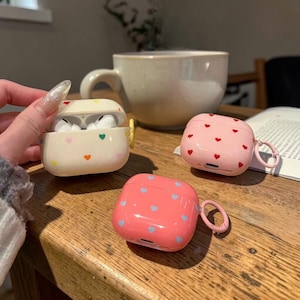 Könnte beinhalten: Drei farbenfrohe AirPod-Hüllen auf einem Holztisch. Eine ist geöffnet und zeigt weiße Ohrhörer. Die Hüllen sind cremefarben, rosa und korallenfarben, jede mit herzförmigen Mustern in verschiedenen Farben. Im Hintergrund steht eine weiße Tasse.