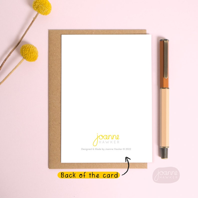 May include: The back of a blank greeting card with a brown envelope. The card has a white background with the text "joanne HAWKER" and "Designed & Made by Joanne Hawker &copy; 2022" printed in black. A brown pen is lying on the right side of the card.