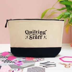 May include: A white canvas zippered pouch with a black base. The pouch has black text that reads "Quilting Stuff" with a stylised needle and thread design.