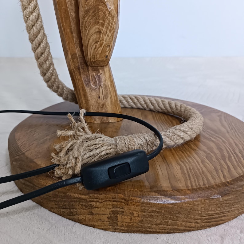 Peut inclure: Gros plan sur un pied de lampe en bois avec un accent de corde naturelle. La lampe pr&eacute;sente une base en bois marron et un support en bois assorti. Un cordon d'alimentation noir avec un interrupteur marche/arr&ecirc;t est visible. La corde est enroul&eacute;e autour de la base et du support.