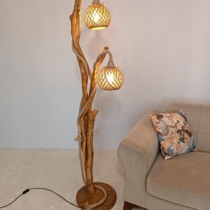 Peut inclure: Un lampadaire rustique avec une structure en bois et deux abat-jours globes tress&eacute;s. La lampe est enroul&eacute;e de corde et poss&egrave;de une base ronde en bois. La lampe est &agrave; c&ocirc;t&eacute; d'un canap&eacute; beige avec un coussin floral.