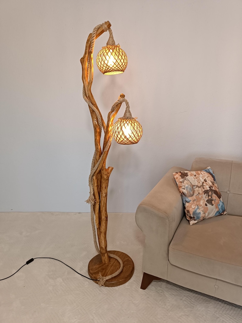 Peut inclure: Un lampadaire rustique avec une structure en bois et deux abat-jours globes tress&eacute;s. La lampe est enroul&eacute;e de corde et poss&egrave;de une base ronde en bois. La lampe est &agrave; c&ocirc;t&eacute; d'un canap&eacute; beige avec un coussin floral.