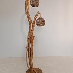 Peut inclure: Lampadaire artisanal avec une base en bois naturel et une structure en forme d'arbre. Deux abat-jours globulaires tiss&eacute;s sont suspendus aux branches, et une corde entoure le tronc. La lampe a une base ronde et un cordon d'alimentation noir.