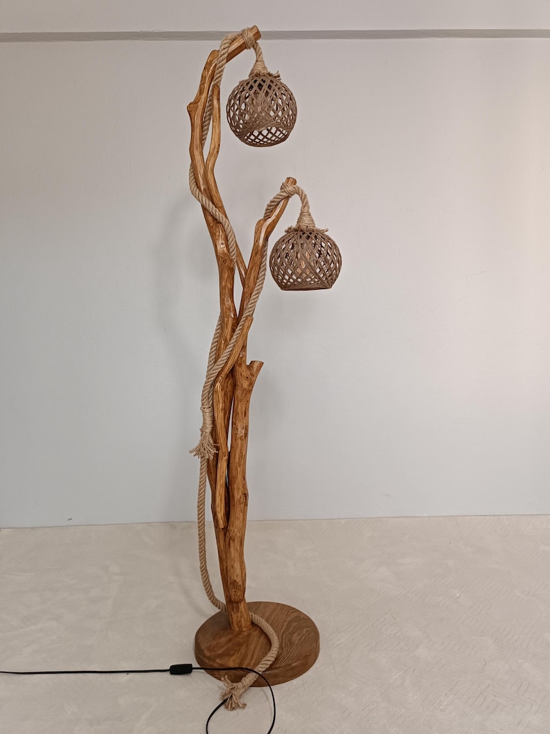 Peut inclure: Lampadaire artisanal avec une base en bois naturel et une structure en forme d'arbre. Deux abat-jours globulaires tiss&eacute;s sont suspendus aux branches, et une corde entoure le tronc. La lampe a une base ronde et un cordon d'alimentation noir.