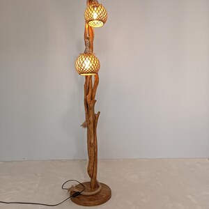 Peut inclure: Lampadaire rustique avec une base et un tronc en bois naturel. Deux abat-jours globes en rotin tress&eacute; sont suspendus en haut, &eacute;clair&eacute;s. La lampe est orn&eacute;e de d&eacute;tails en corde et d'un c&acirc;ble d'alimentation noir. Hauteur: environ 180 cm.