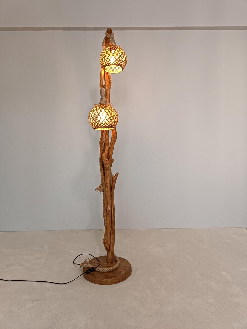 Peut inclure: Lampadaire rustique avec une base et un tronc en bois naturel. Deux abat-jours globes en rotin tress&eacute; sont suspendus en haut, &eacute;clair&eacute;s. La lampe est orn&eacute;e de d&eacute;tails en corde et d'un c&acirc;ble d'alimentation noir. Hauteur: environ 180 cm.